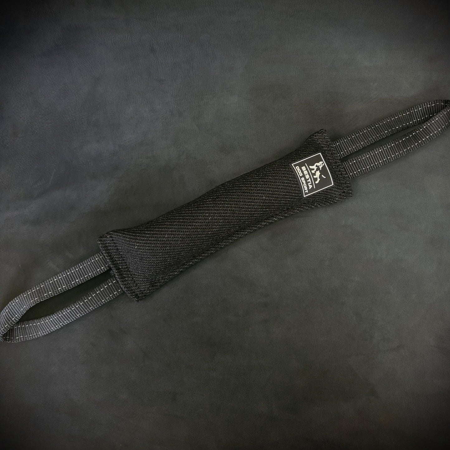 Close-up of a black Bestia Dog Sport Bite Tug for dog training, showing the durable woven fabric stretched out, the white brand logo, and two heavy-duty nylon handles on a dark background.