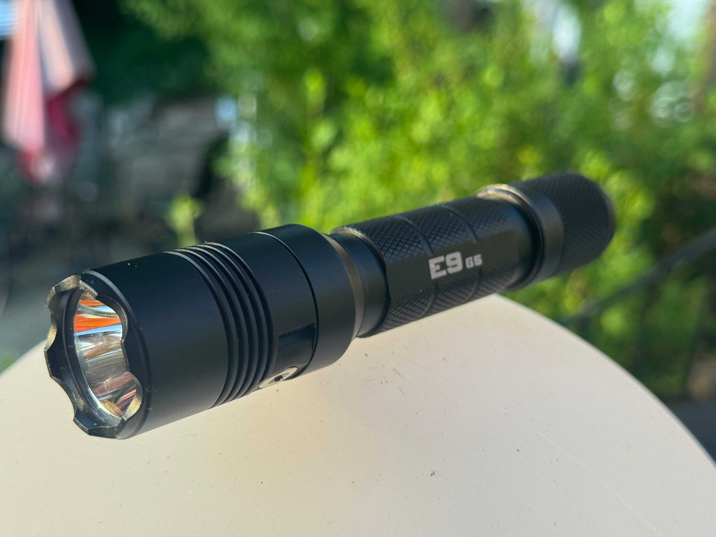 Close-up of a Powertac E9 G5 tactical flashlight resting on a white surface with a blurred green garden background.
