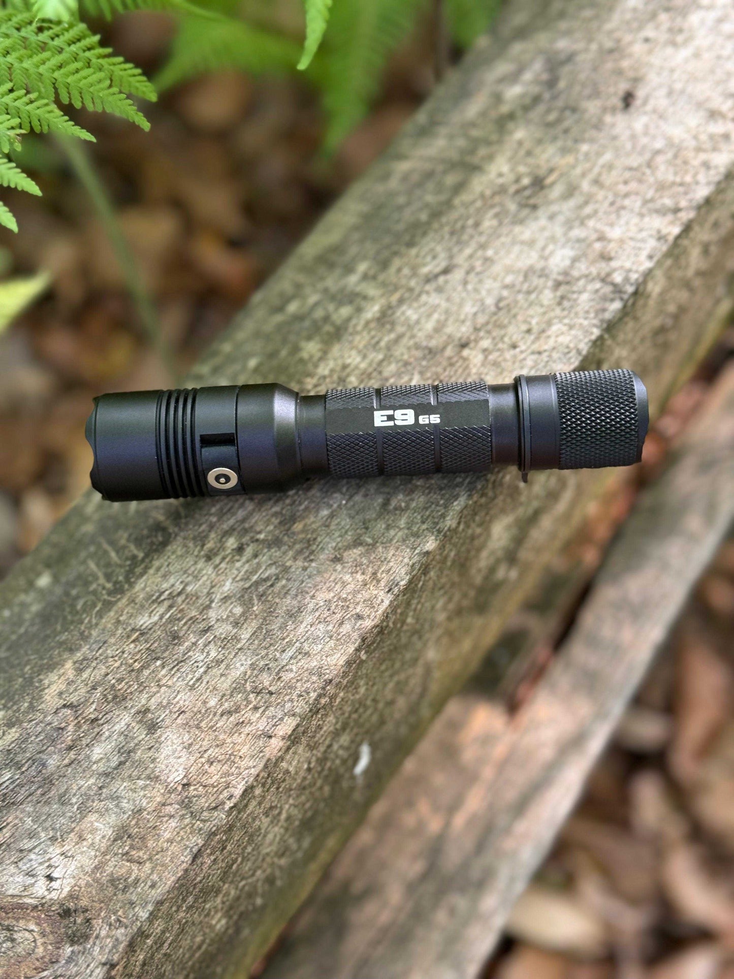 Powertac E9 G5 tactical flashlight resting on a weathered wooden beam in a forest setting with green ferns in the background.