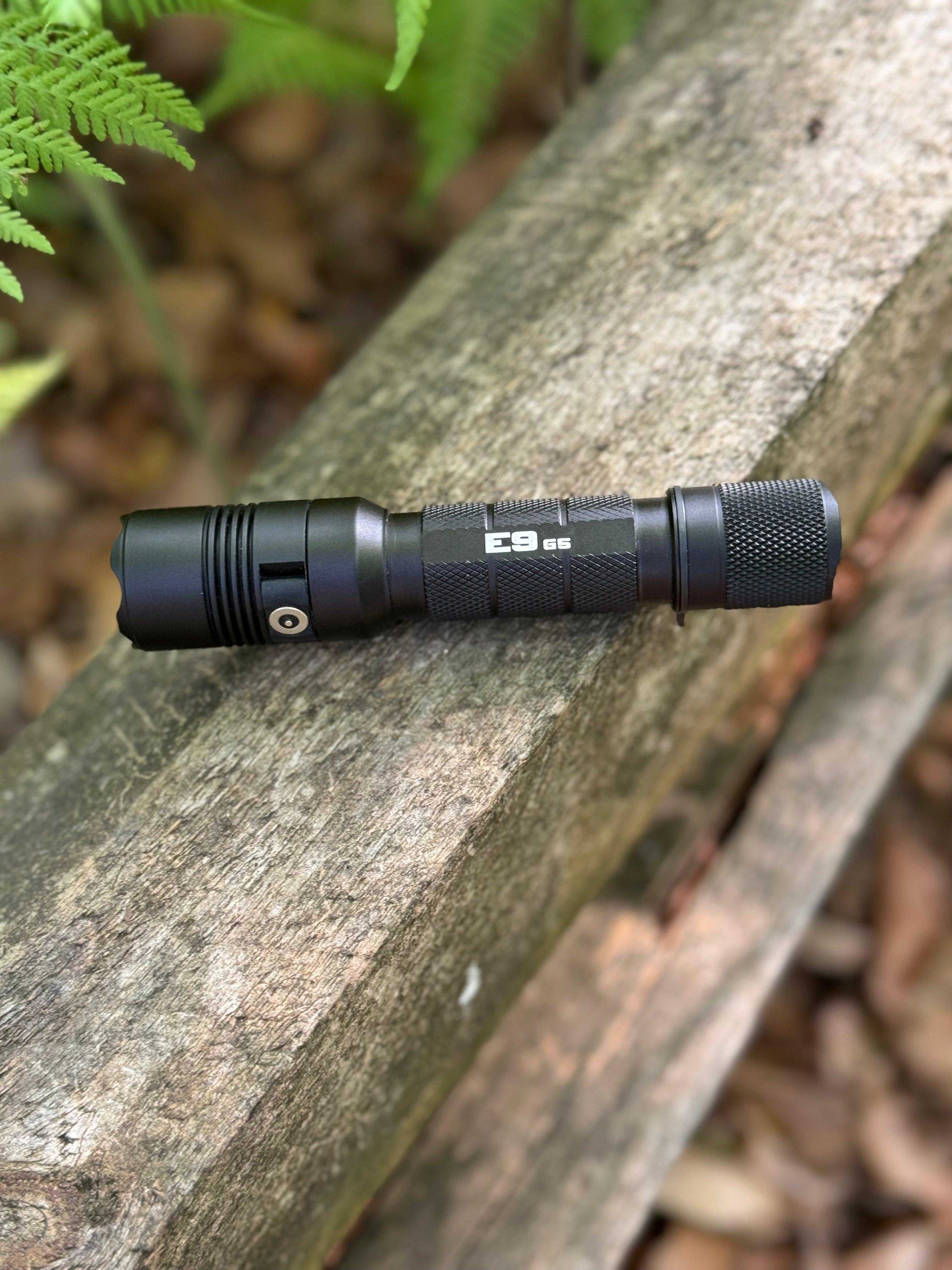 Powertac E9 G5 tactical flashlight resting on a weathered wooden beam in a forest setting with green ferns in the background.