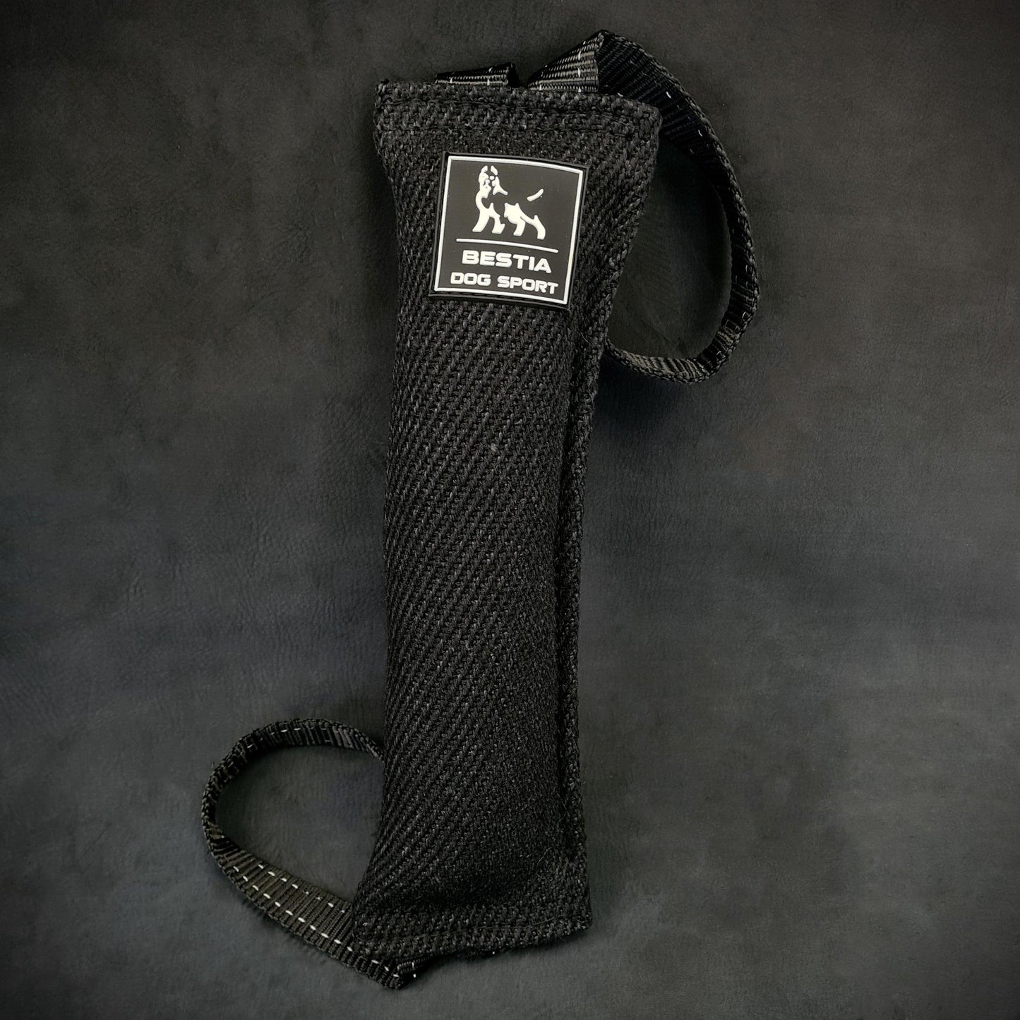 Close-up of a black Bestia Dog Sport Bite Tug for dog training, showing the durable woven fabric, the white brand logo, and two heavy-duty nylon handles on a dark background.