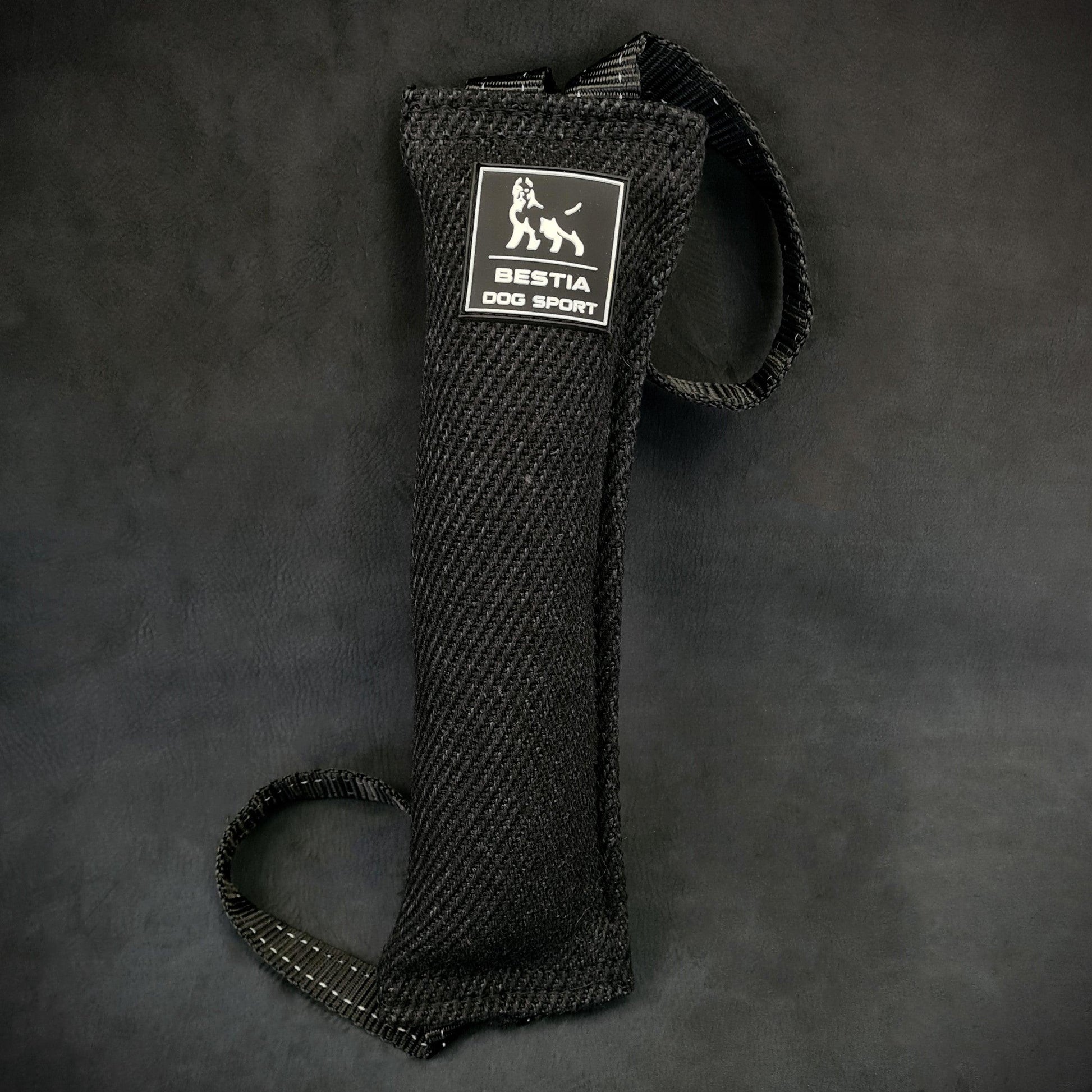Close-up of a black Bestia Dog Sport Bite Tug for dog training, showing the durable woven fabric, the white brand logo, and two heavy-duty nylon handles on a dark background.