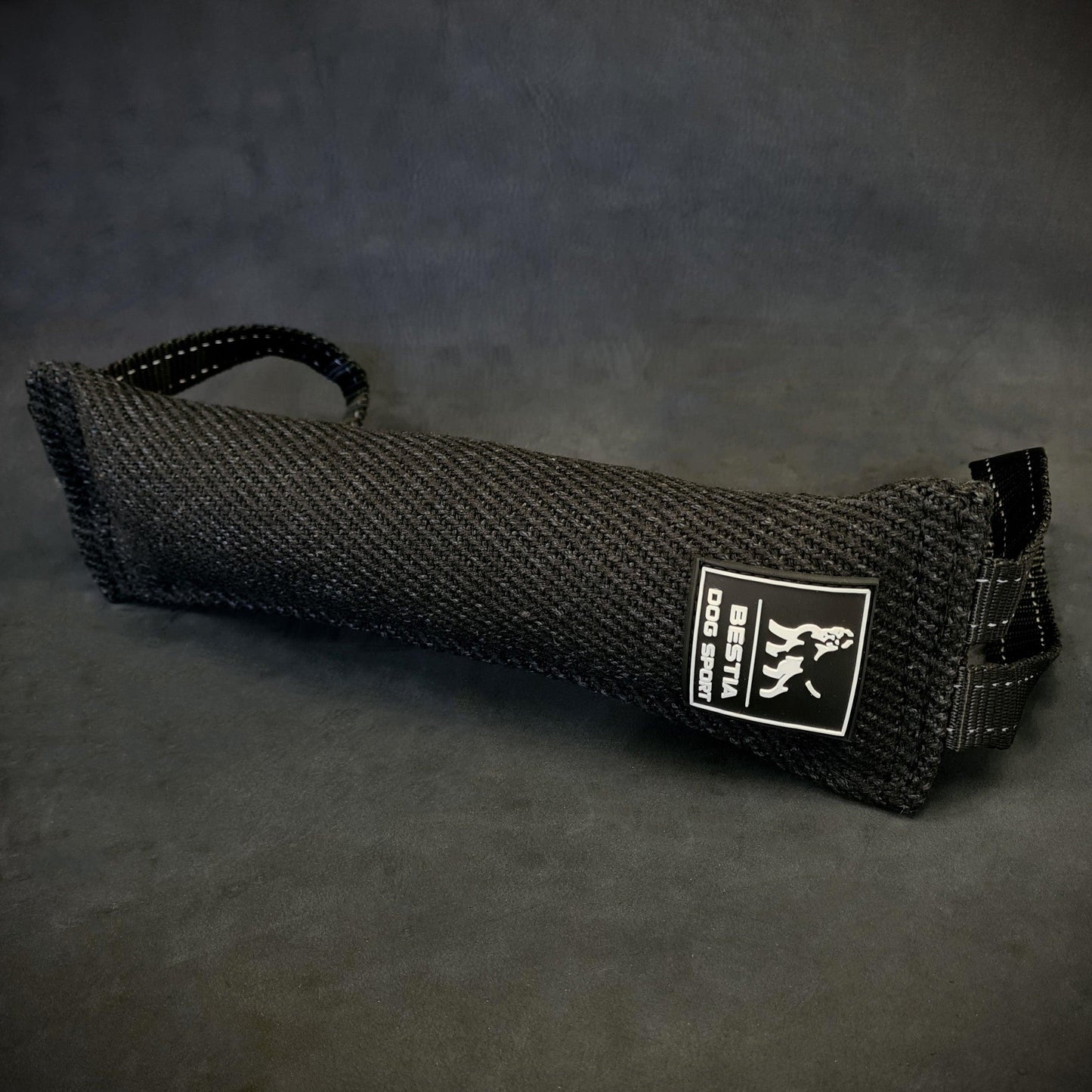 Close-up of a black Bestia Dog Sport Bite Tug for dog training, showing the durable woven fabric, the white brand logo, and two heavy-duty nylon handles on a dark background.
