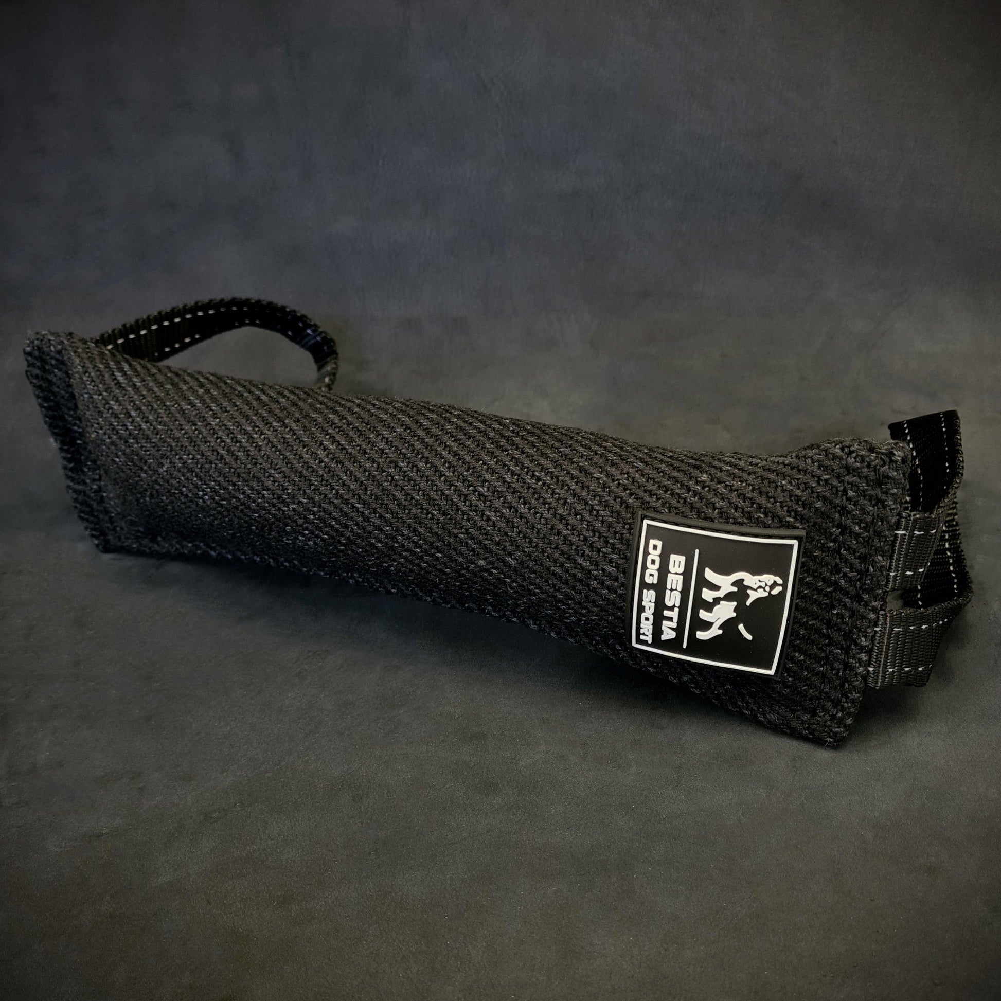 Close-up of a black Bestia Dog Sport Bite Tug for dog training, showing the durable woven fabric, the white brand logo, and two heavy-duty nylon handles on a dark background.