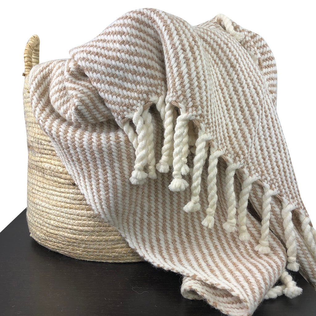 Beige and brown striped blanket with tassels draped over a woven basket on a black surface.