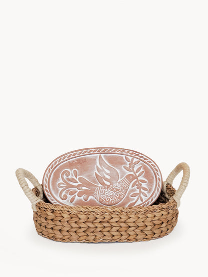 A KORISSA bread warmer set with a decorative bird-engraved terracotta stone leaning inside a hand-woven seagrass basket against a white background.
