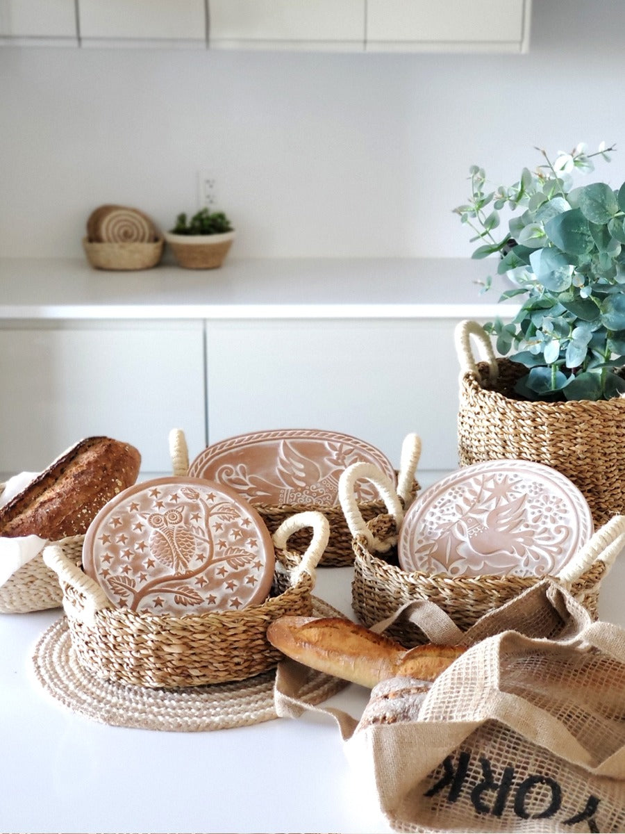 Handcrafted terracotta bread warmers in woven seagrass baskets styled on a bright kitchen counter with fresh bread.