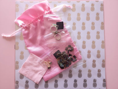 A pink satin drawstring jewelry pouch shown with rings, earrings, and a necklace placed inside small, clear protective plastic sleeves with black velcro closures, resting on a decorative pineapple-patterned background.