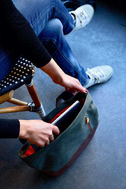 A person placing a slim laptop into the open main compartment of an olive green Canopy Verde 3-in-1 bag, showcasing its vibrant orange lining and capacity as a functional work or school bag.