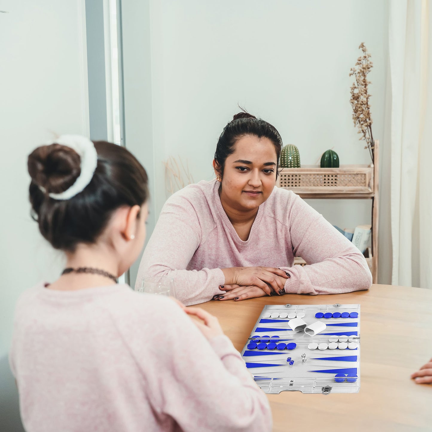 Two people playing a premium lucite backgammon set at a table in a bright modern home, showcasing the elegant acrylic design in use